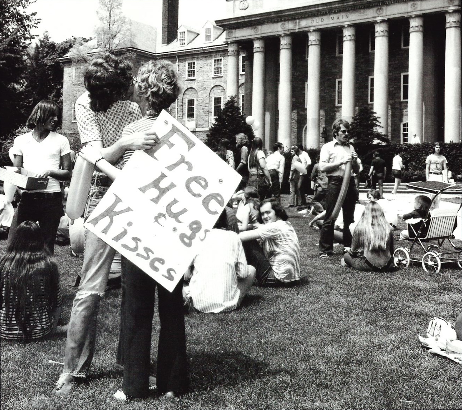 black and white photo of a woman holding a sign that reads free hugs and kisses while kissing a man with other student in the background on the Old Main lawn by Penn State Archives