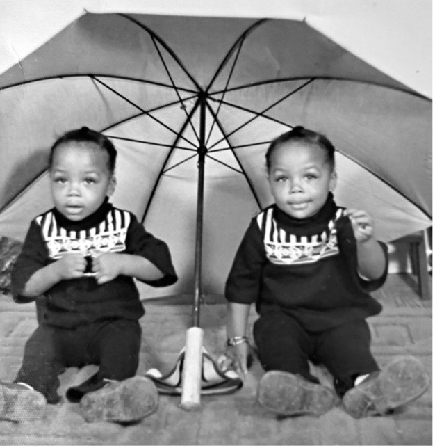 Delana Wardlaw and Elana McDonald as babies in matching outfits under a large umbrella, courtesy