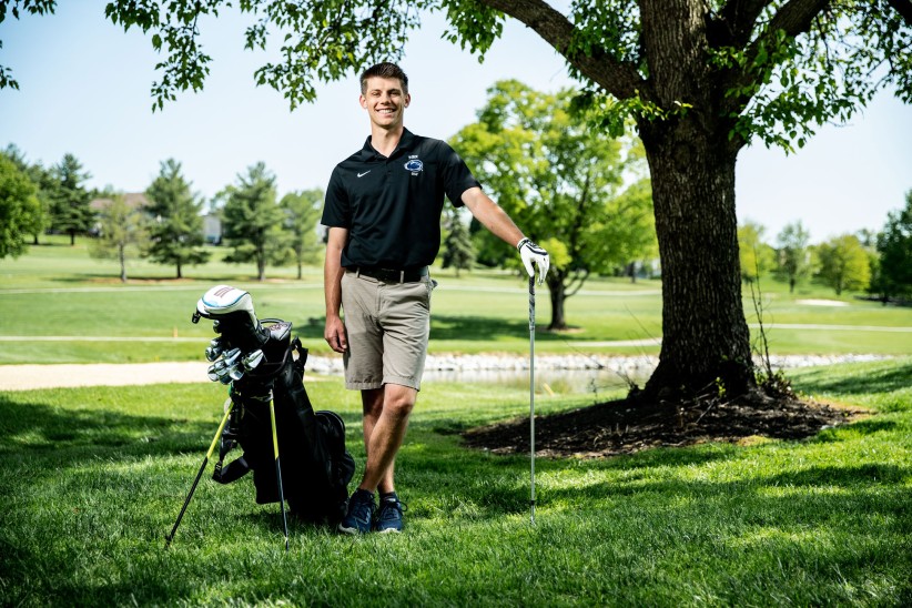 Logan Snyder and wearing a Penn State golf polo with clubs under a tree by Cardoni