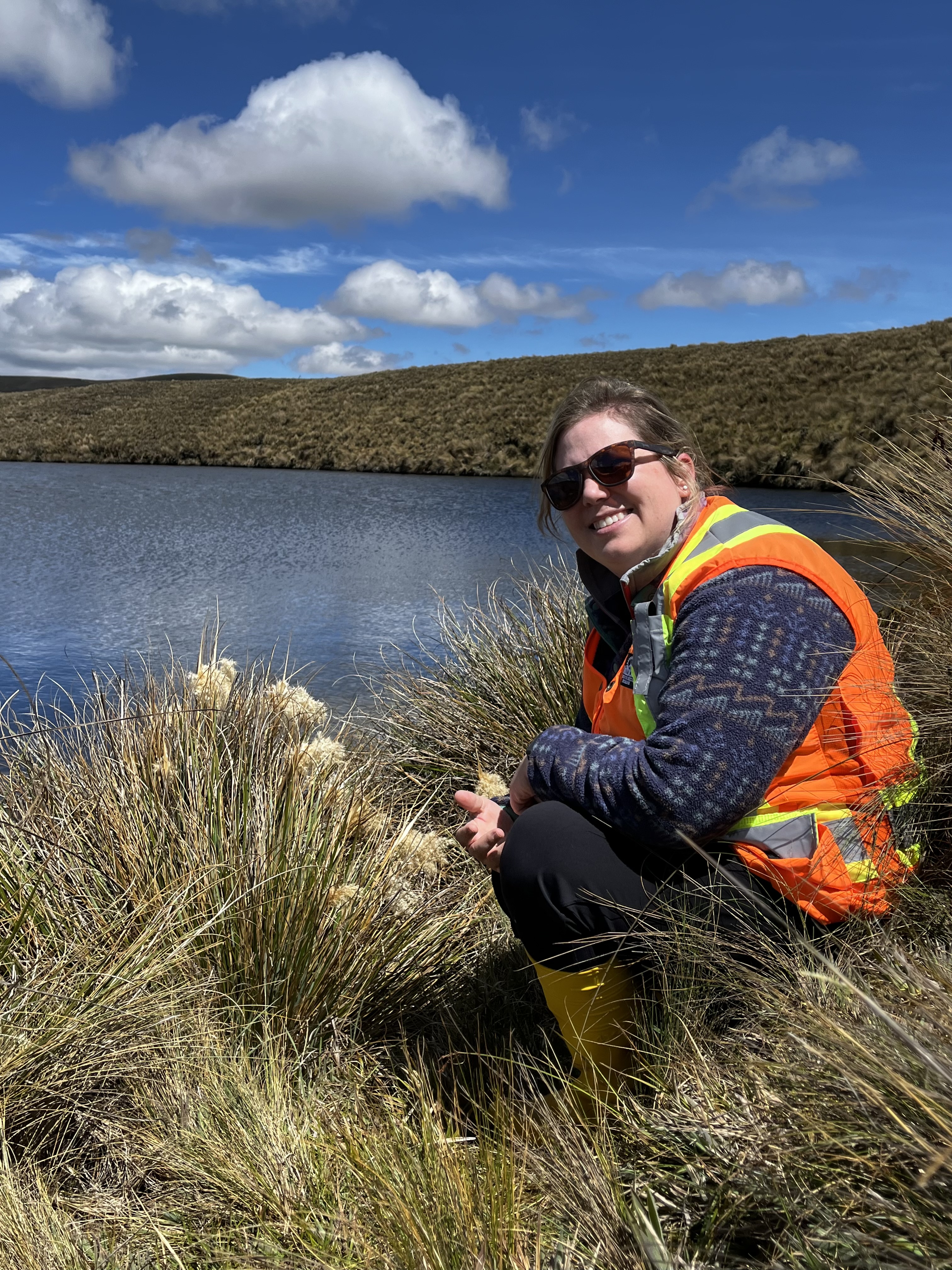 Guifarro kneeling in tall grass next to a body of water with mountains in the background, courtesy