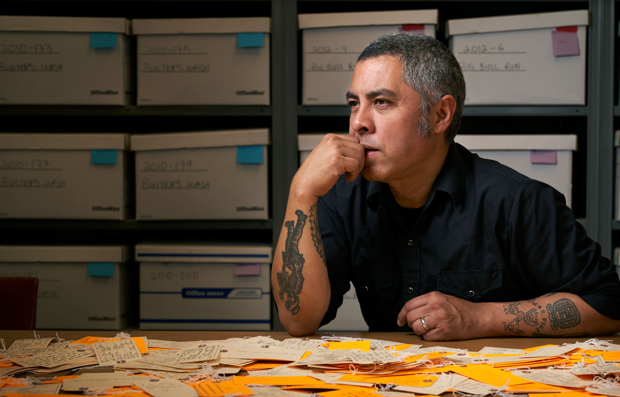 De Leon seated at desk with an array of luggage tags on the table in front of him and assorted backpacks on shelves behind him, photo by Gregg Segal