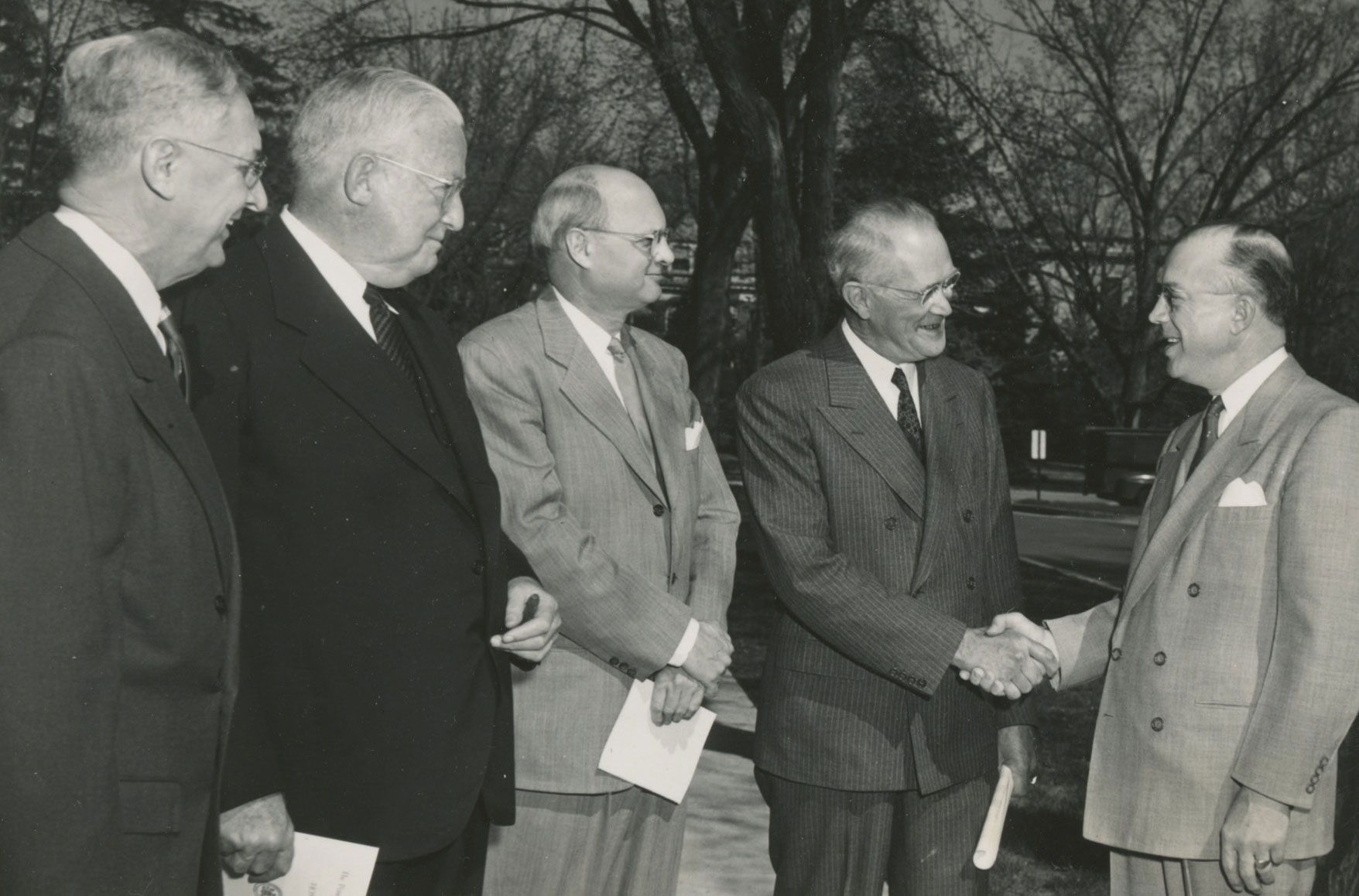 black and white photo of four men shaking hands outside, by PSAA