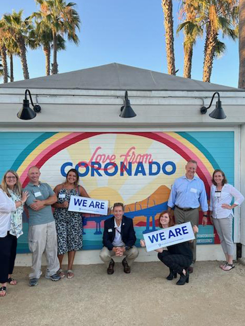 photo of alums holding We Are signs standing in front of a mural that says Love from Coronado with palm trees in background, courtesy Alumni Career Services