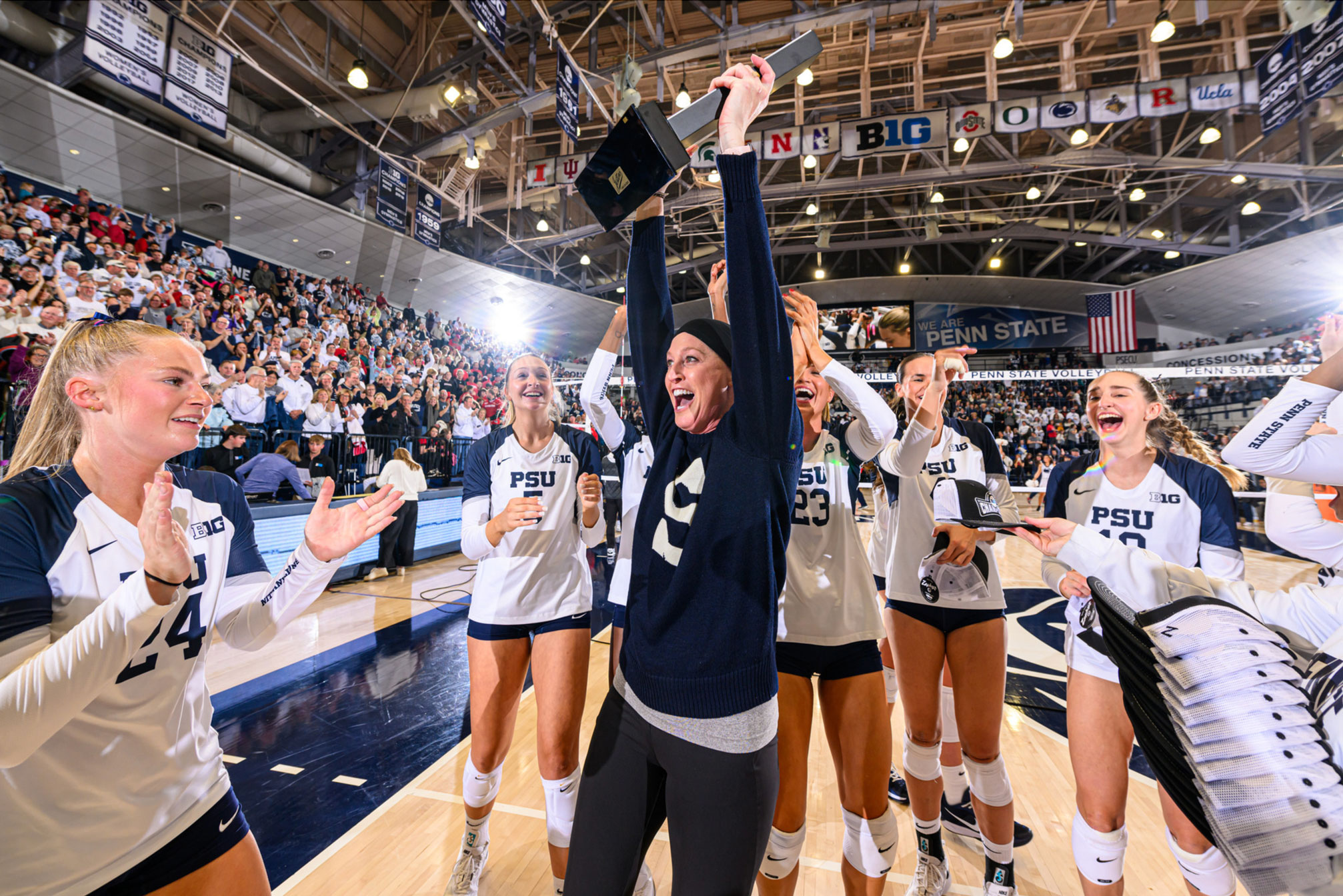 Nittany Lion women's volleyball coach Katie Schumacher-Cawley hoists Big 10 trophy overhead while players cheer, photo by Penn State Athletics