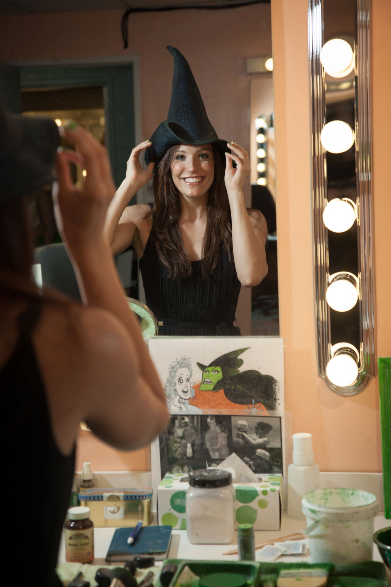 Caroline Bowman looking in a backstage mirror to try on a witch hat, photo by Michael Lavine
