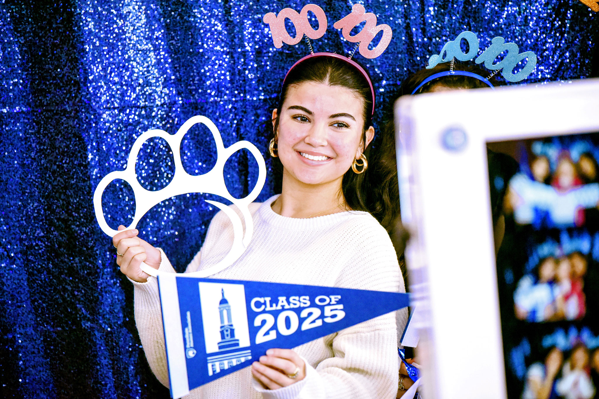 photo of a student holding a class of 2025 banner and a white Nittany Lion paw and wearing a pink 100 headband during the 100 Days 'til Graduation event, by Penn State Alumni Association