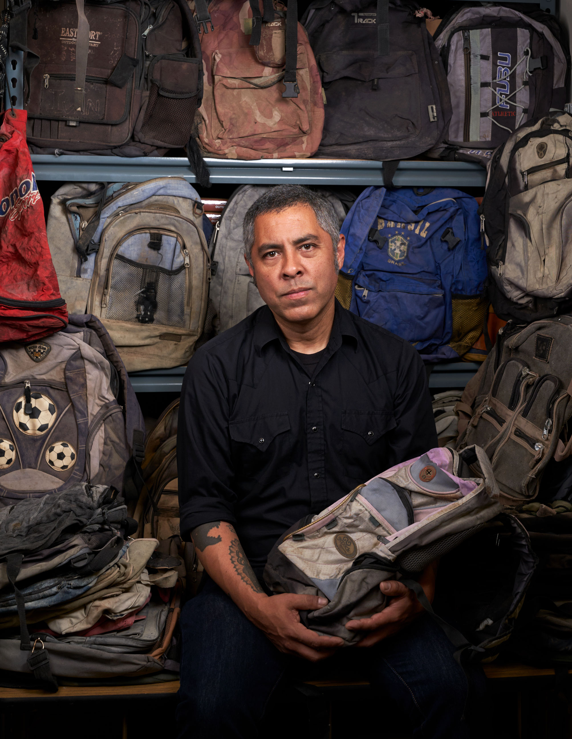 De Leon holding a backpack with array of backpacks on shelves behind him, photo by Gregg Segal