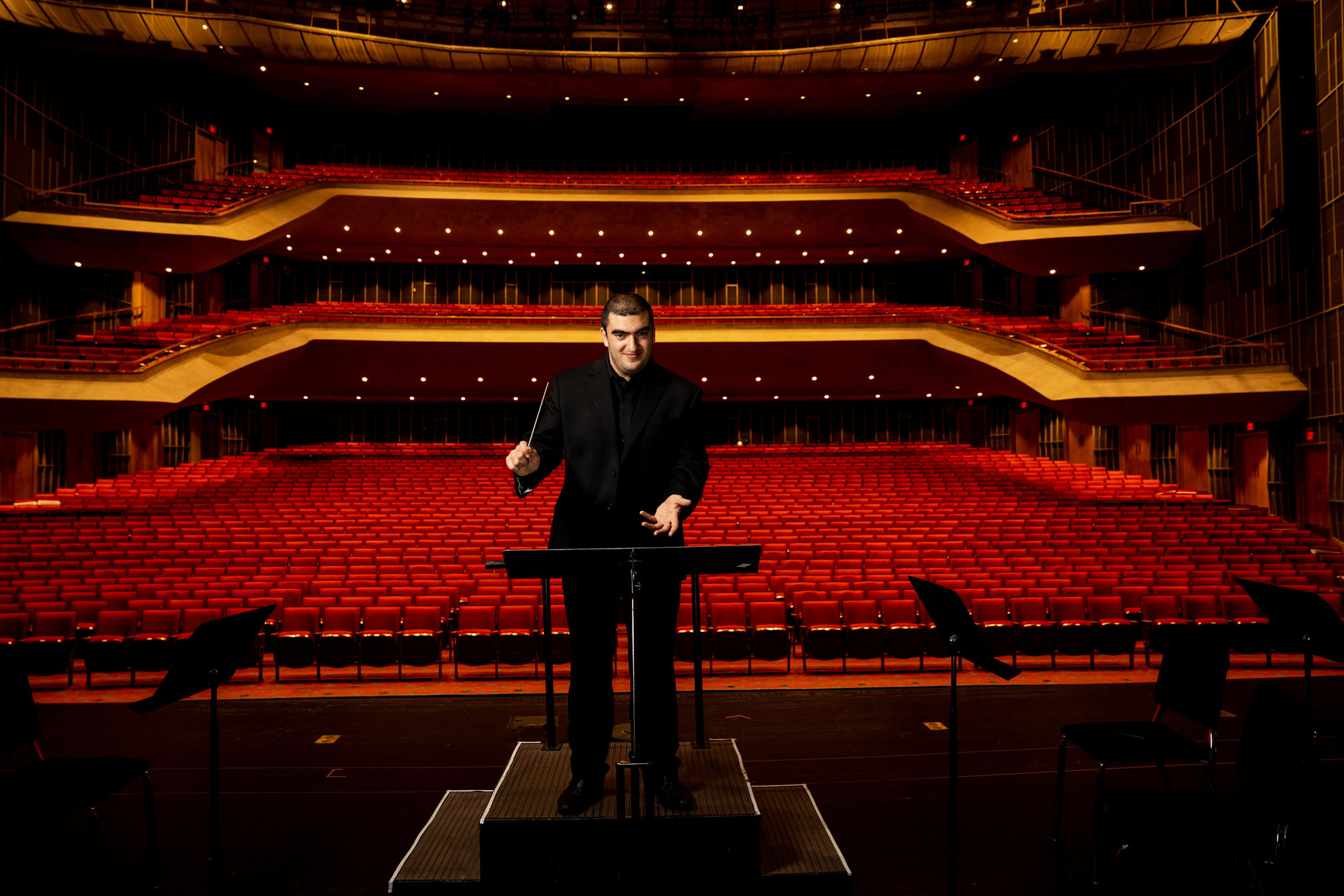 Amran conducting in an empty theater with hundreds of empty red chairs behind him by Cardoni