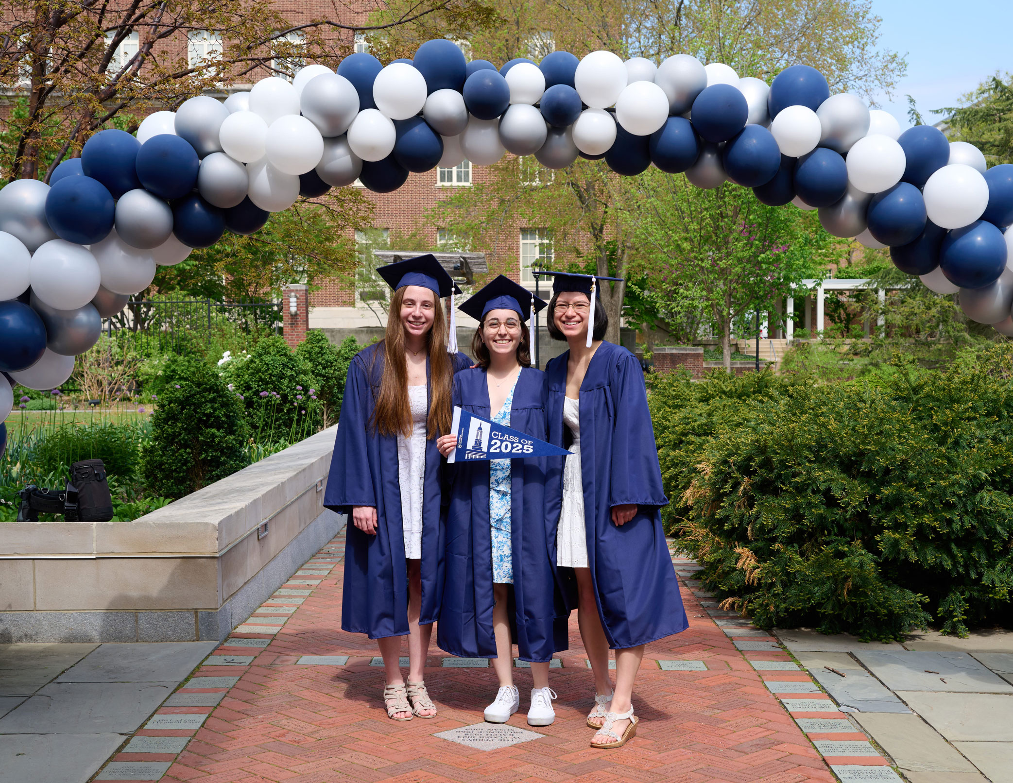 photo of three graduates in caps and gowns under blue and white balloon arch outside Hintz by Penn State Alumni Association