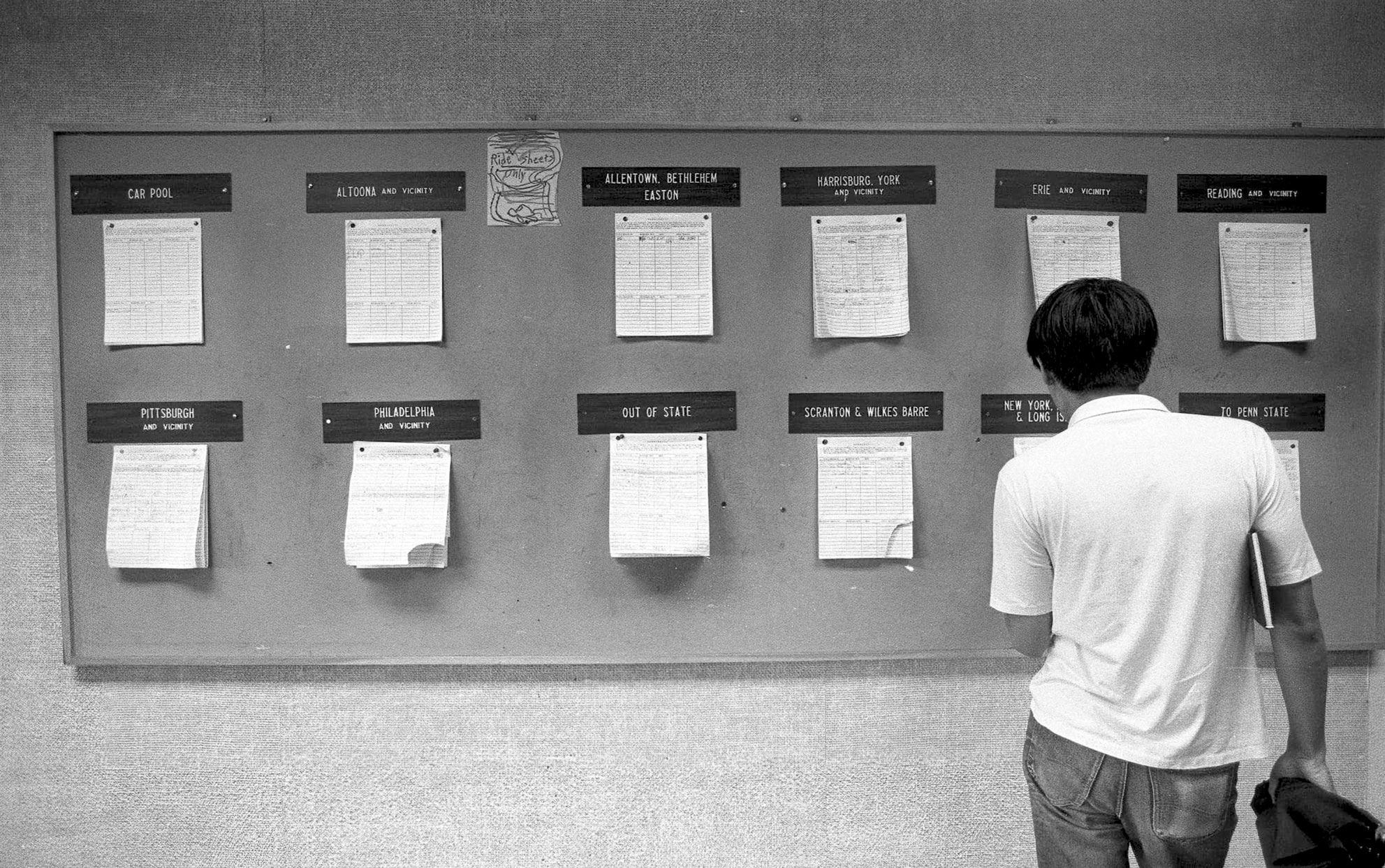 black and white photo of a student standing in front of a large bulletin board with multiple sheets of paper tacked to it by Pat Little