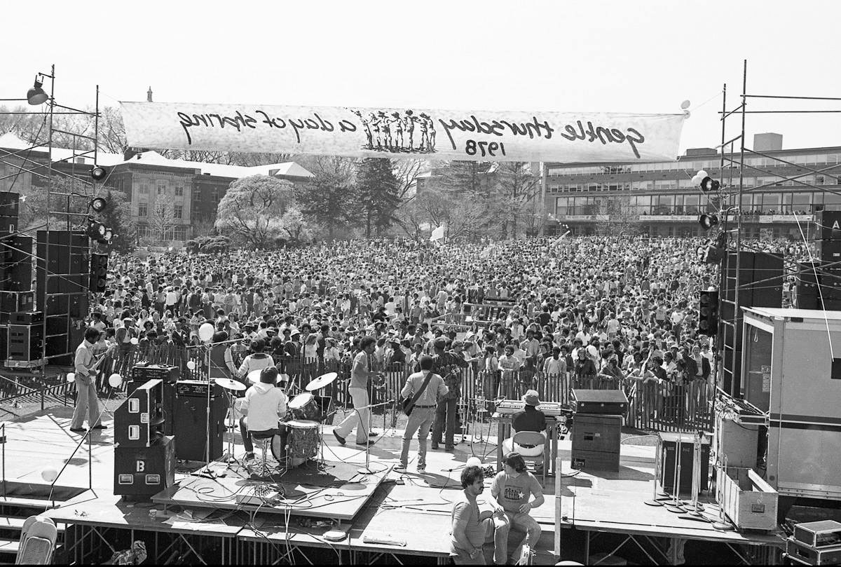 black and white photo of a large stage outside with a band playing to a large crowd by Pat Little