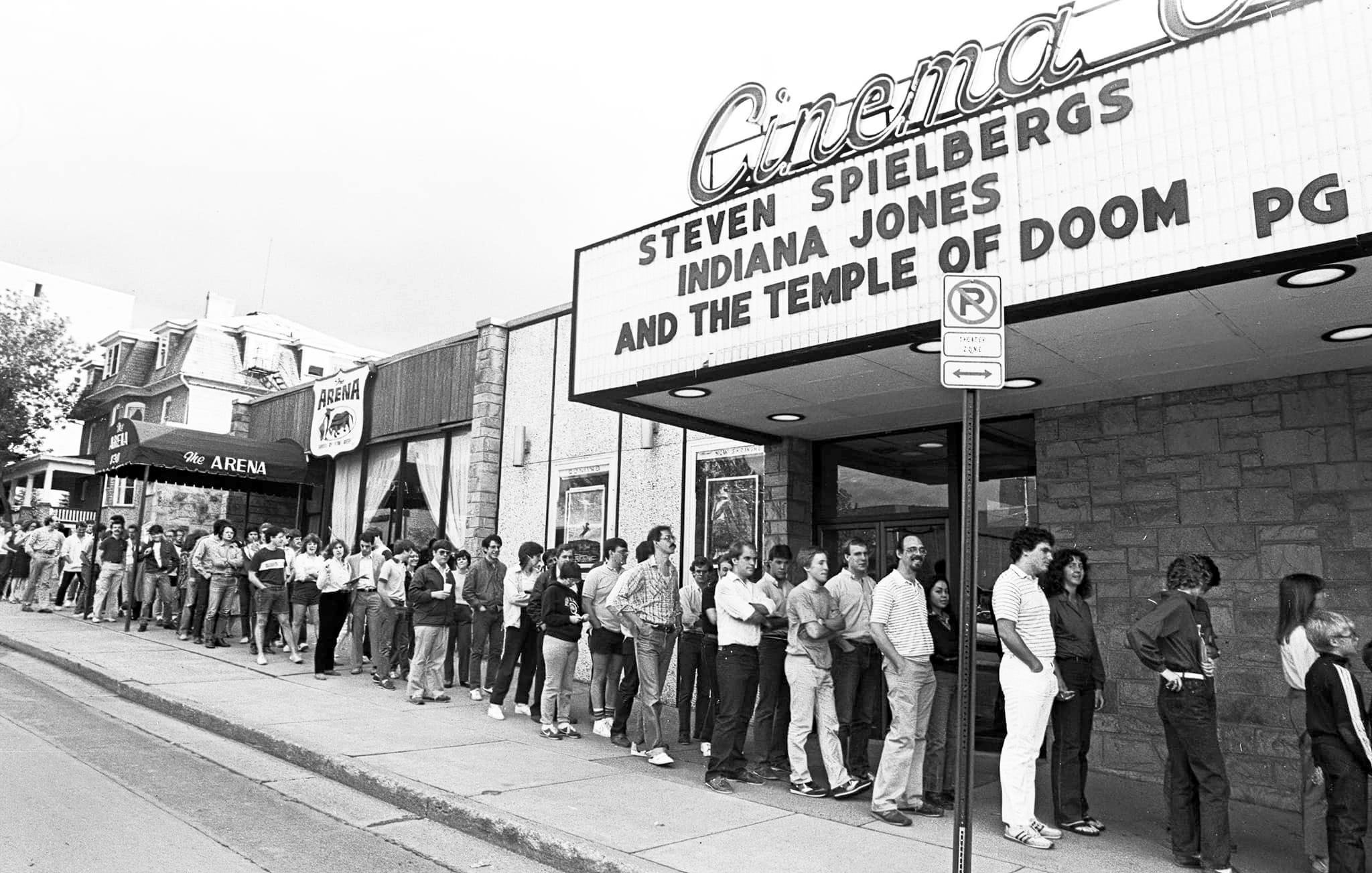 black and white photo of a long line of people outside a movie theater advertising "Steven Spielbergs Indian Jones and the Temple of Doom" by Pat Little