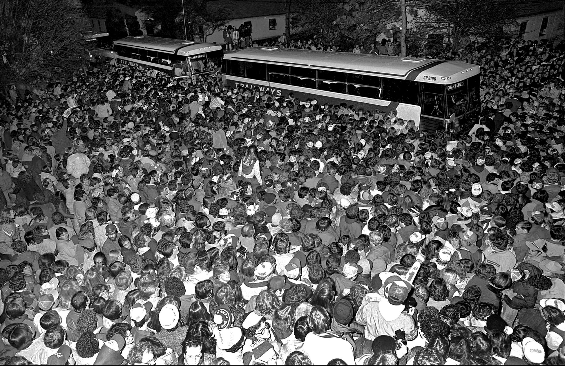black and white photo of a large crowd of people greeting the arrival of a caravan of tour busses by Pat Little