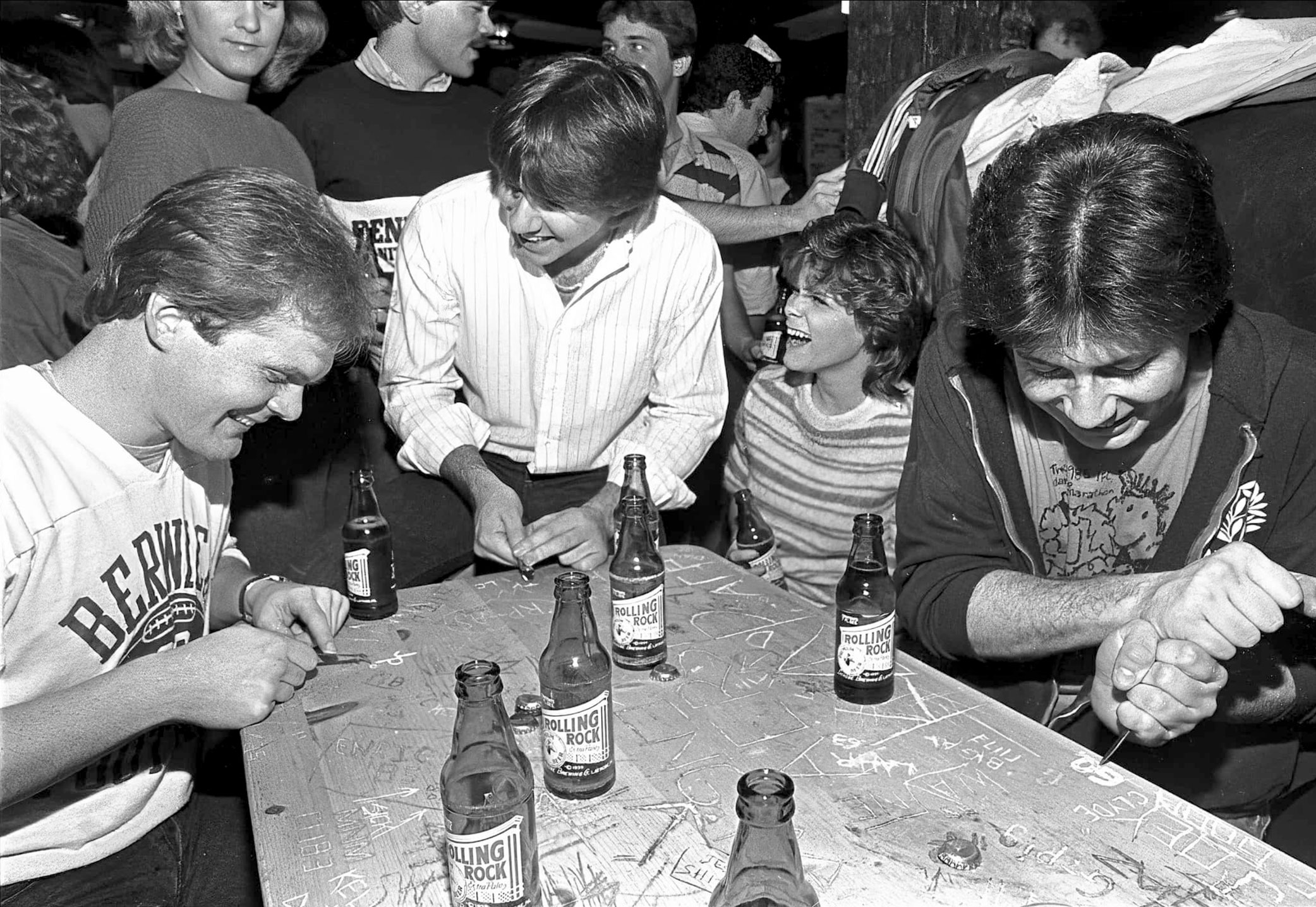 black and white photo of a group of four people at The Rathskeller carving names into a wooden table on which sits multiple Rolling Rock pony bottles by Pat Little