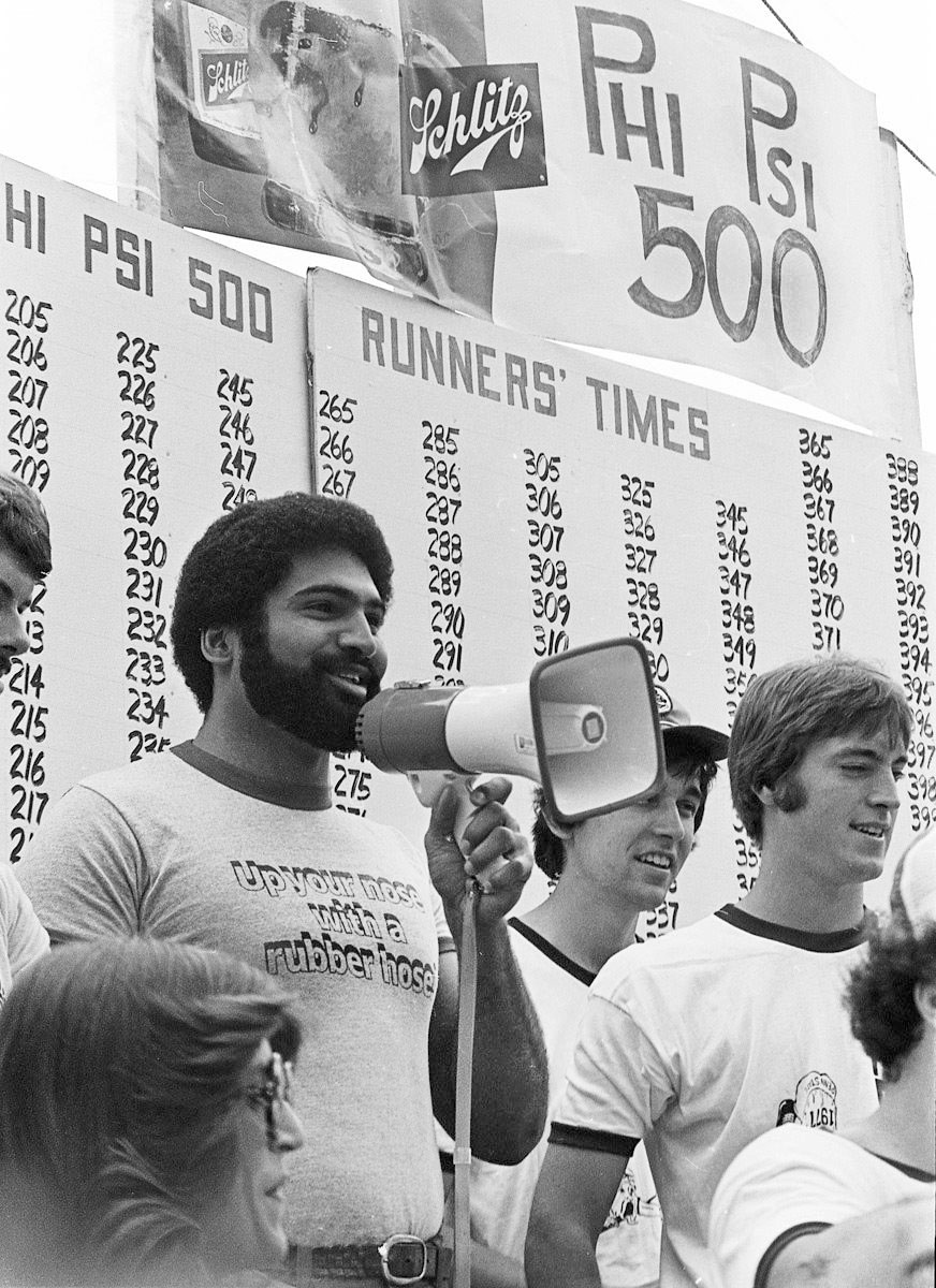 black and white photo of Franco Harris speaking into a megaphone standing before a board with runners' times by Pat Little