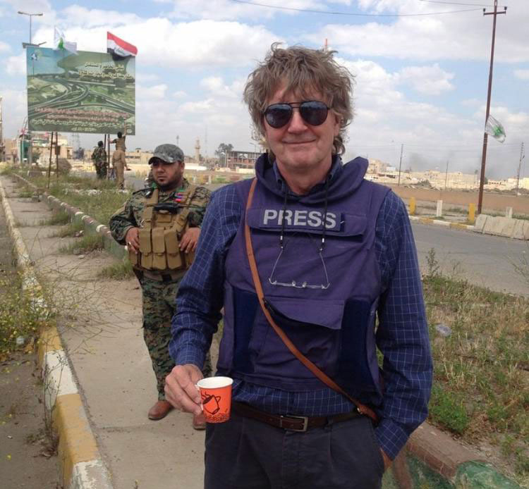 Norland wearing a press vest and holding a coffee cup alongside a road in Iraq, courtesy