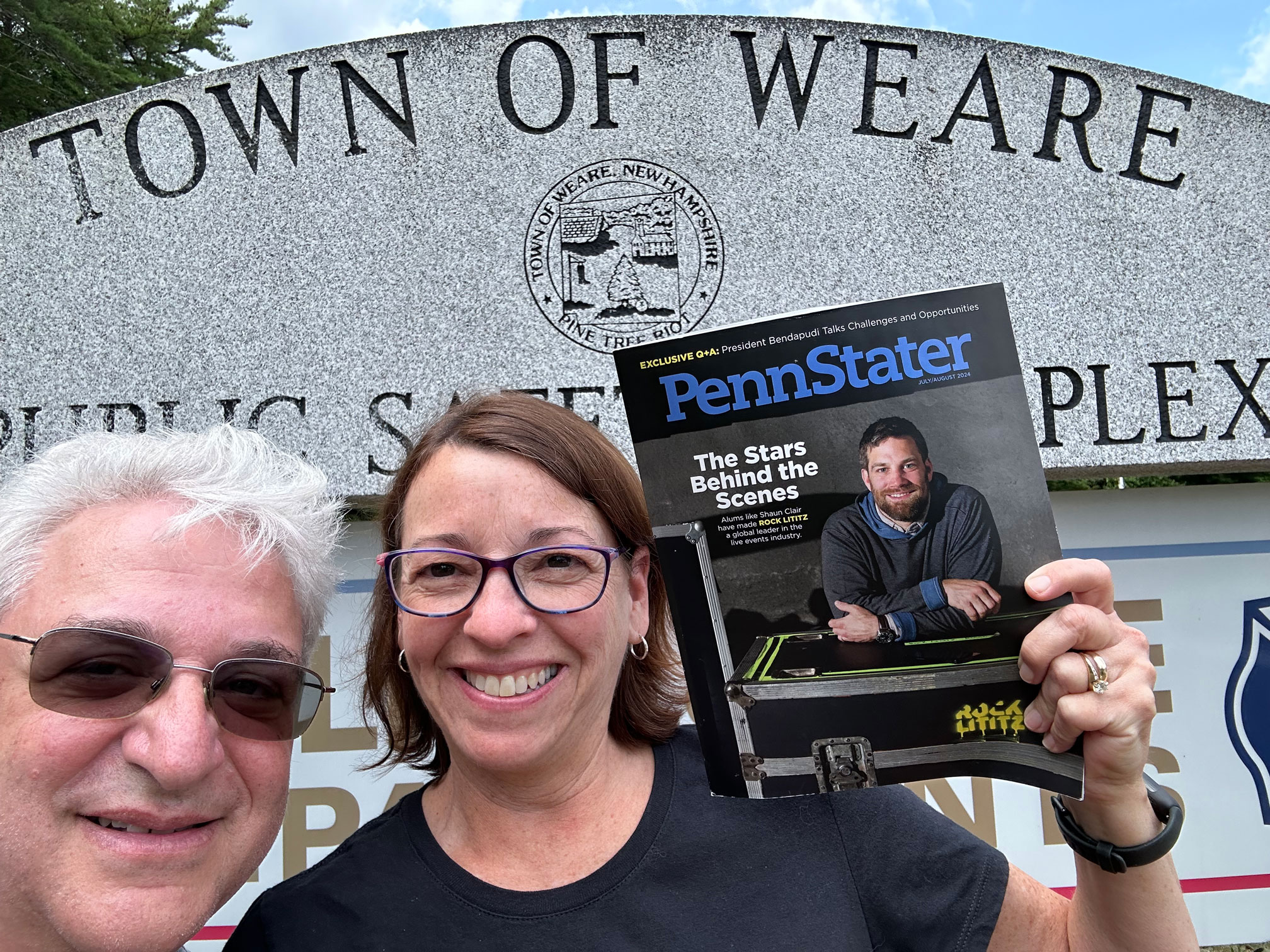 two alums holding up a copy of the magazine in front of a stone sign welcoming visitors to the town of WEARE, courtesy