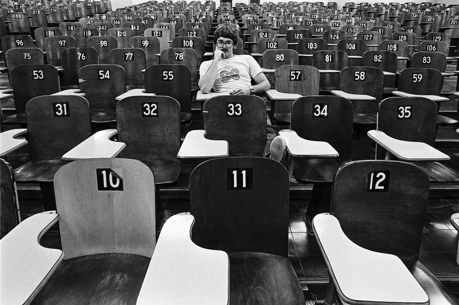 black and white photo of Pat Little sitting in an empty lecture hall with numbered wooden seats by Pat Little