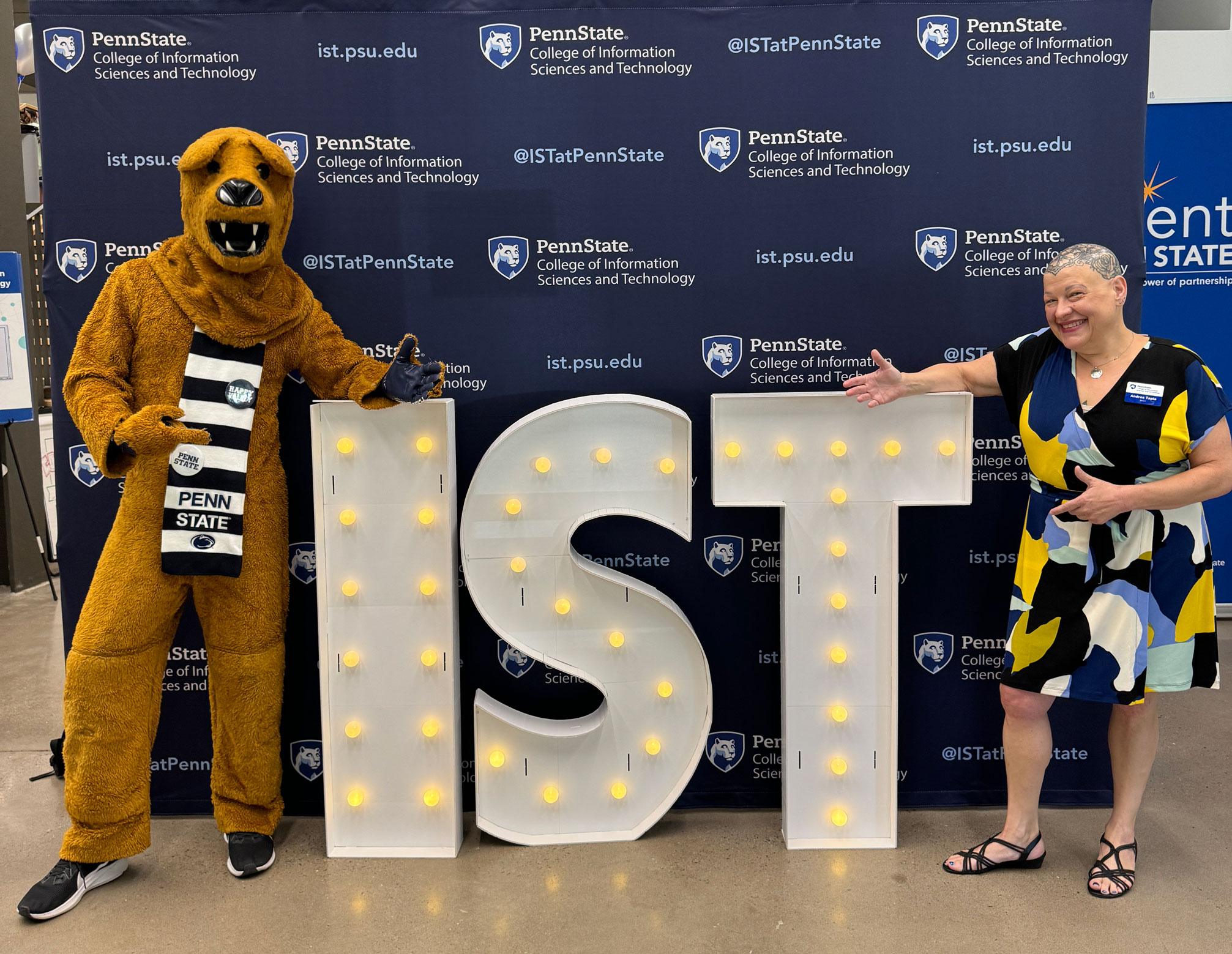 Nittany Lion and Tapia gesturing to a large lit up IST sign by Penn State