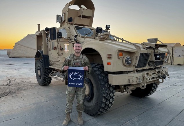 photo of Senior airman Alexander Dugan holding a Penn State flag in front of an army vehicle, courtesy