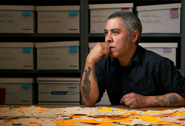 De Leon seated at desk with an array of luggage tags on the table in front of him and assorted backpacks on shelves behind him, photo by Gregg Segal