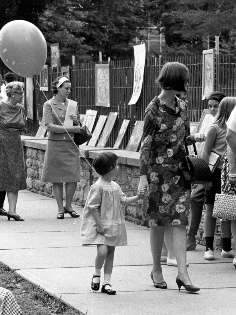Black and white photo of a woman and child holding hands walking down a street during Arts Fest, by Penn State Archives