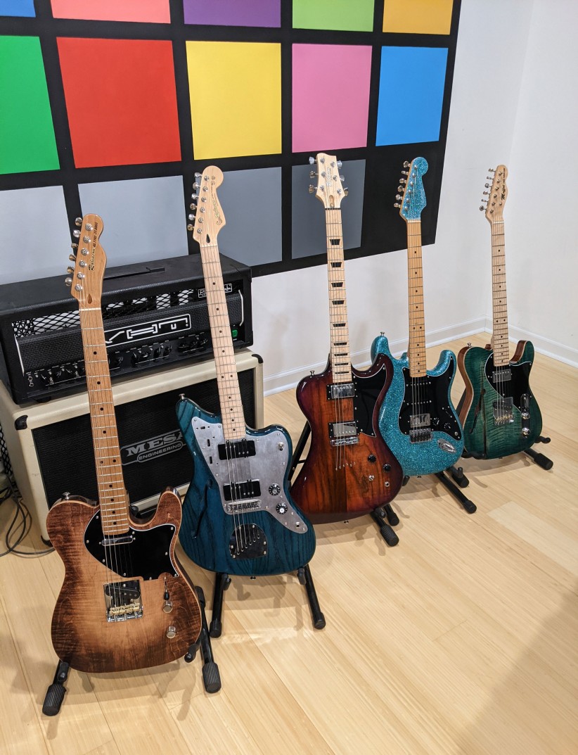 five colorful electric guitars on stands in front of an amp with a color block wall behind them, courtesy Anthony Robinson