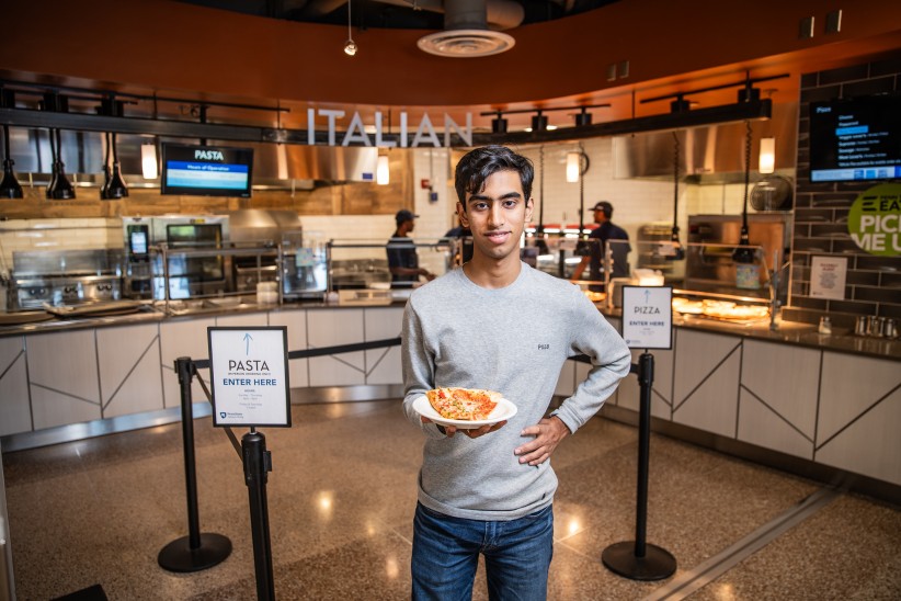 photo of Ryan Nair holding a plate with a slice of pizza in front of a campus dining spot by Cardoni
