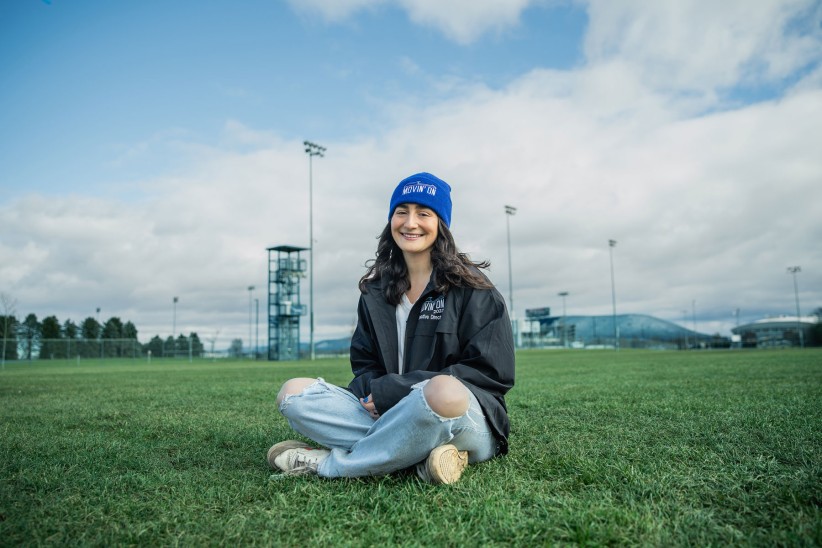Photo of Sofia Moussan sitting cross-legged on green grass wearing a blue Movin On hat by Cardoni