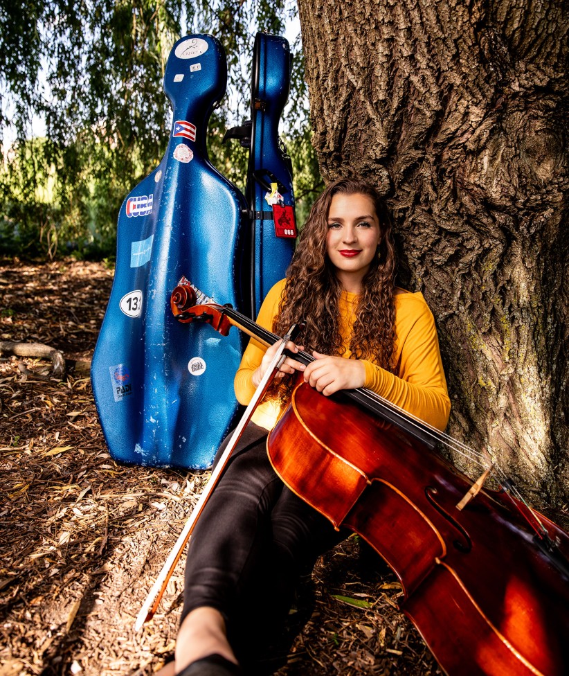 Ruth Stokes sitting outside against a tree with her cello, photo by Cardoni