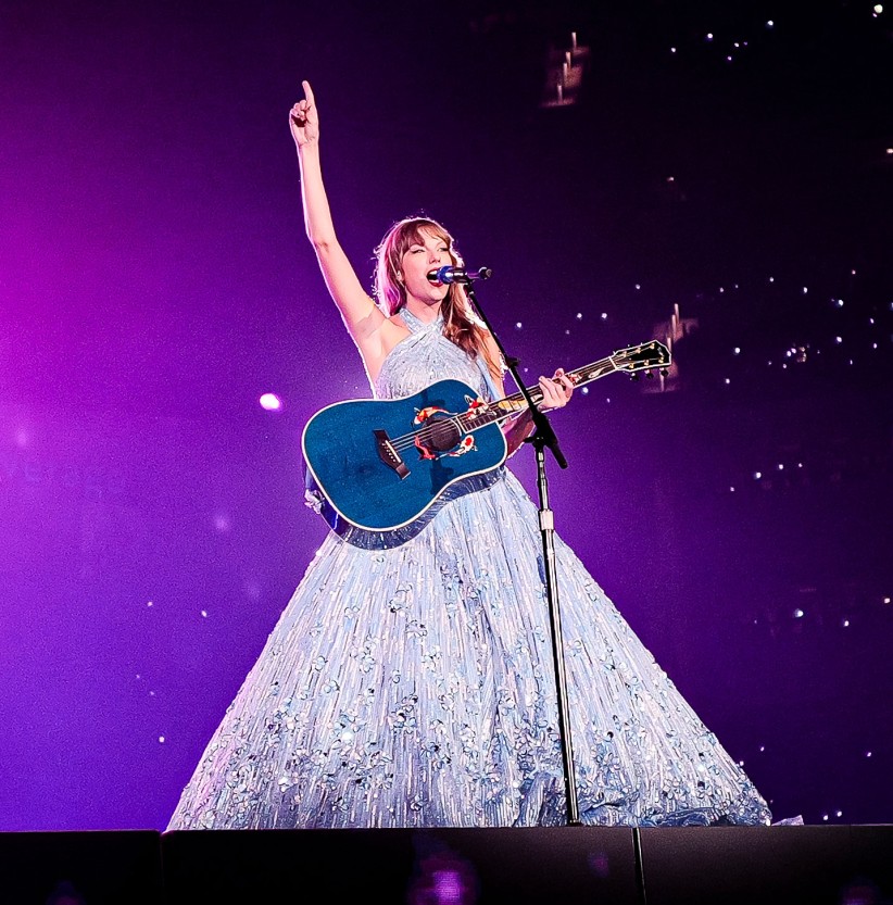 photo of Taylor Swift onstage in white gown holding blue guitar with arm raised overhead and background lit in purple by Paolo Villanueva