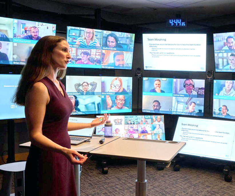 woman standing at a desk speaking to multiple screens with people on Zoom, photo by Steve Tressler