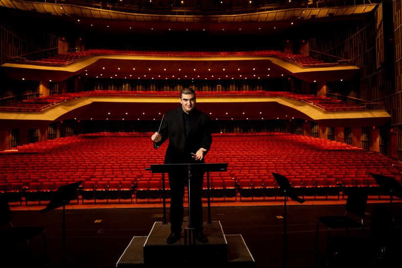 Amron conducting on stage with an enormous empty theater of red seats behind him by Cardoni