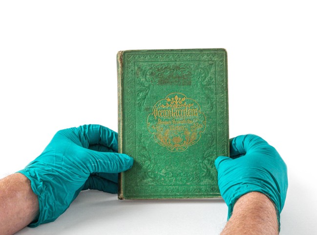 Photo of two gloved hands holding an old book with an emerald green cover and gold lettering. The emerald green dye contained arsenic and requires PPE when handling. Photo by Nick Sloff '92 A&A
