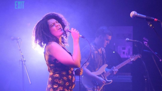 Kat Leverenz singing on a purple-lit stage, courtesy