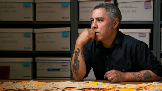 De Leon seated at desk with an array of luggage tags on the table in front of him and assorted backpacks on shelves behind him, photo by Gregg Segal