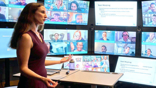 woman standing at a desk speaking to multiple screens with people on Zoom, photo by Steve Tressler