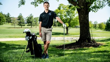 Logan Snyder and wearing a Penn State golf polo with clubs under a tree by Cardoni