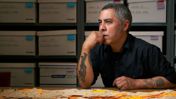 De Leon seated at desk with an array of luggage tags on the table in front of him and assorted backpacks on shelves behind him, photo by Gregg Segal