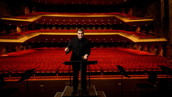 Amron conducting on stage with an enormous empty theater of red seats behind him by Cardoni
